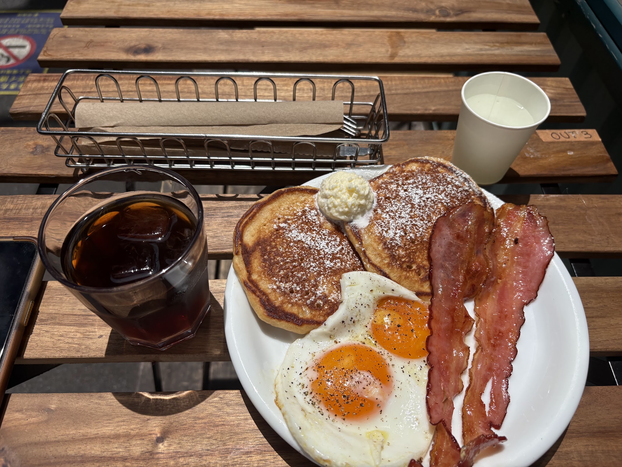 木製テーブルにパンケーキ・ベーコン・目玉焼きのプレートとアイスコーヒーグラス、屋外テラス席での食事シーン