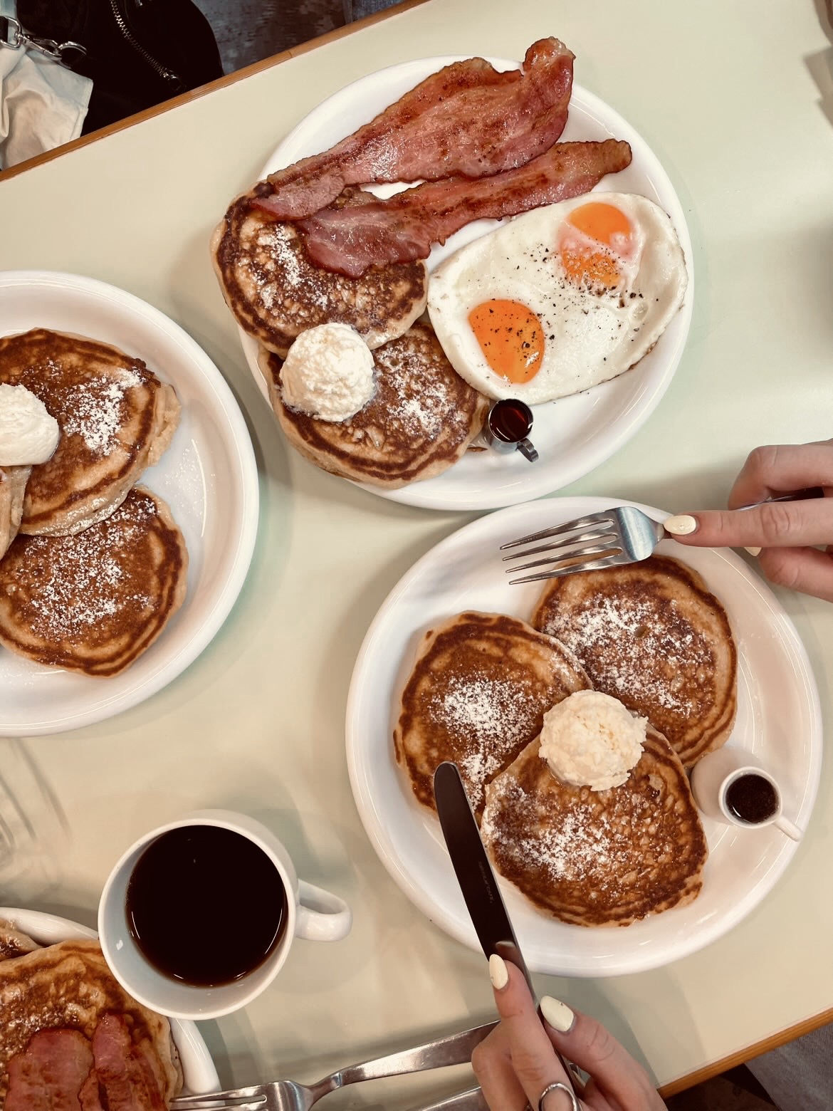 白い丸皿に重なったパンケーキ数枚・目玉焼き2個・ベーコン2枚と、手前のコーヒーカップおよびアイスコーヒーグラスを木製テーブルの上から撮影した俯瞰カット