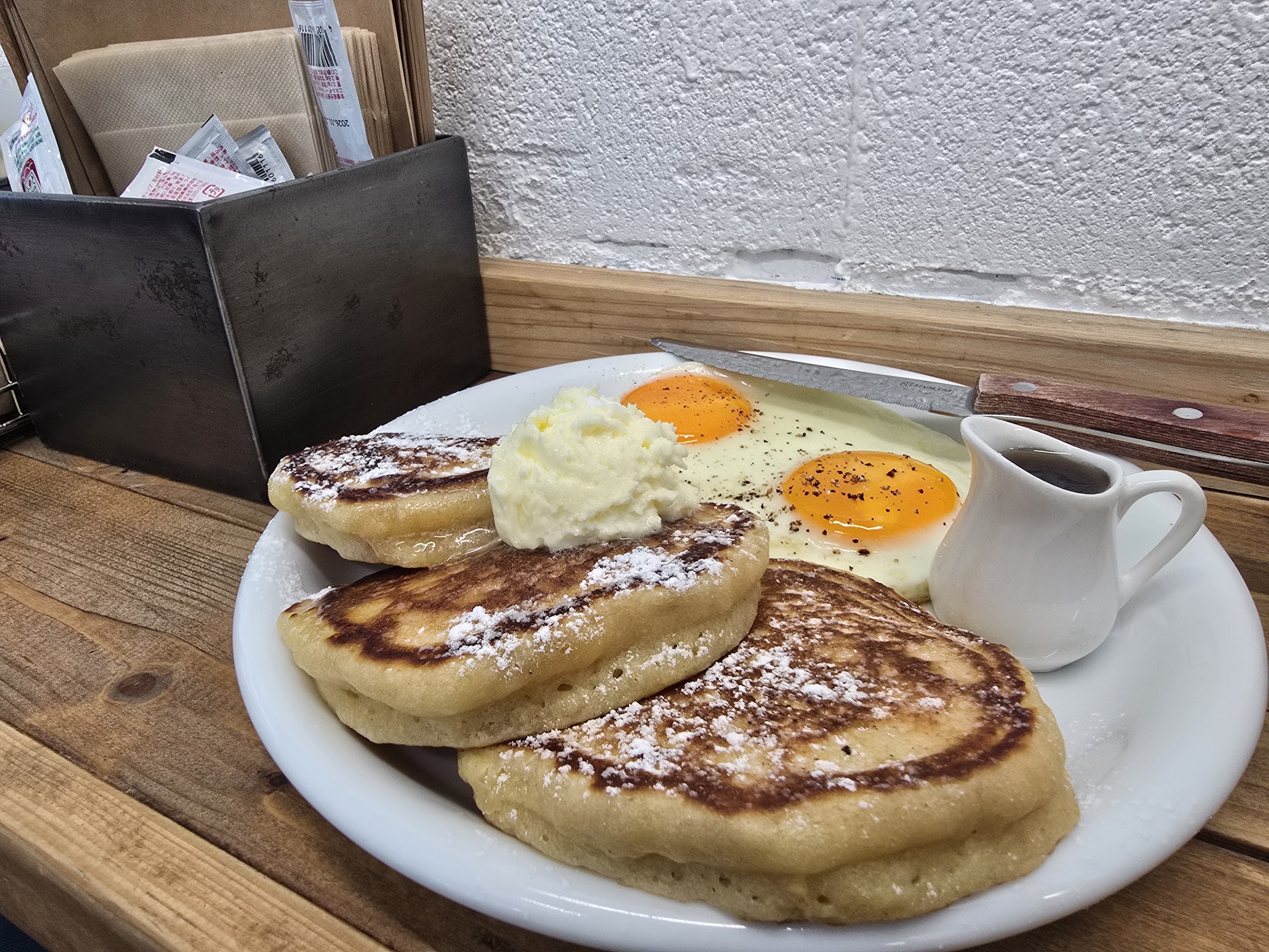 白い楕円皿に盛られた厚みのあるパンケーキ3枚・ホイップクリーム・目玉焼き2個とシロップポット、木製テーブルの上のクローズアップ